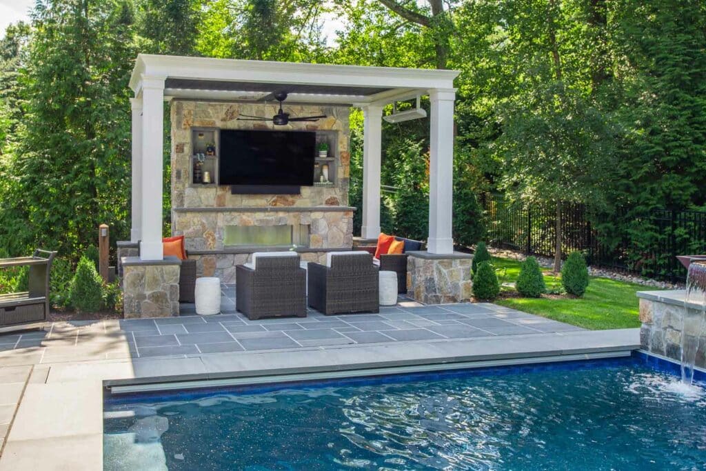Outdoor stone media wall featuring a TV, fireplace and seating by a pool under a white pergola. By Harmony Design Group.