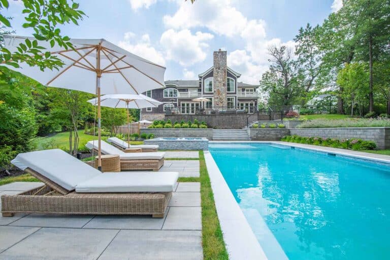 Blue pool, lounge chairs, umbrellas on patio fronting a dark house and garden under a bright sky by Harmony Design Group.