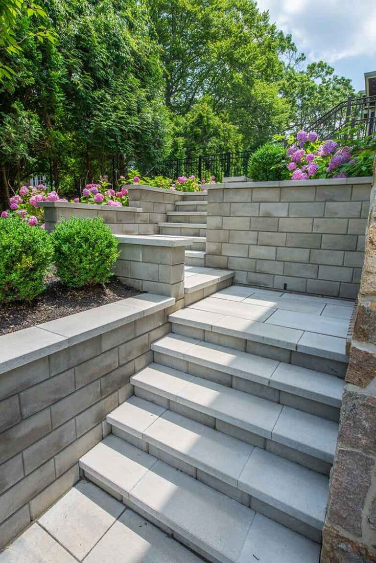 Stone steps ascend between tiered retaining walls with pink hydrangeas and green shrubs, under a sunny sky. By Harmony Design Group