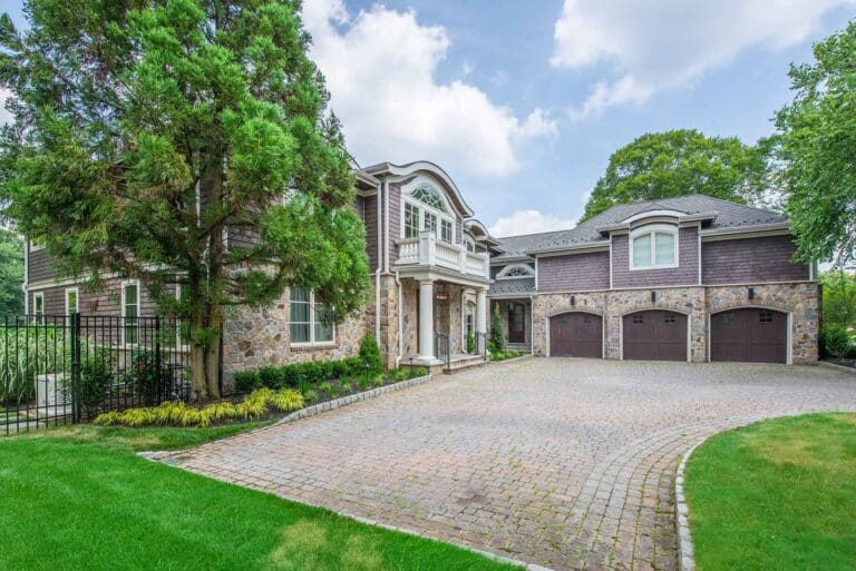 Large stone and shingle house with balcony and 3 garages, fronted by a paved driveway and lush lawn by Harmony Design Group.