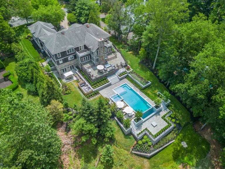 Large house with terraced patios and multi-level pool, surrounded by lush greenery. By Harmony Design Group.