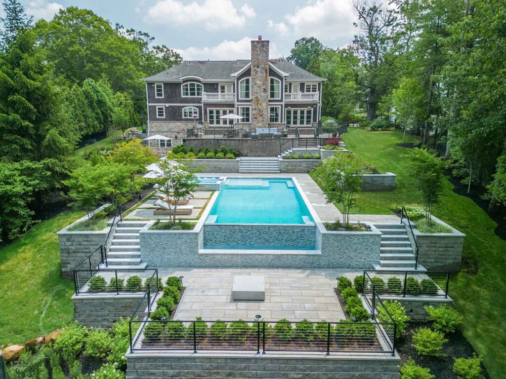 Multi-story house overlooking terraced backyard with an infinity pool, hot tub, patios, and lush landscaping. By Harmony Design Group