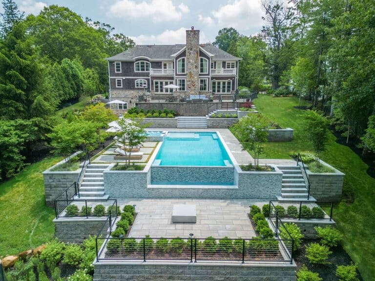 Multi-story house overlooking terraced backyard with an infinity pool, hot tub, patios, and lush landscaping. By Harmony Design Group