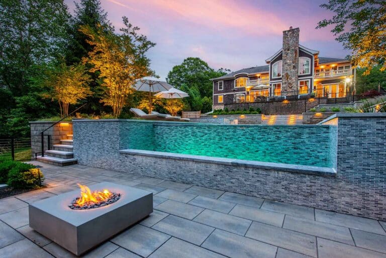 Fire pit burning on a paved patio beside an illuminated infinity pool, with a large house in the background at dusk. By Harmony Design Group.
