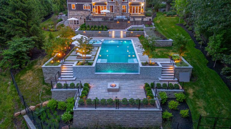 Multi-level backyard showcasing an illuminated pool, spa, and fire pit, leading to a stone house by Harmony Design Group.