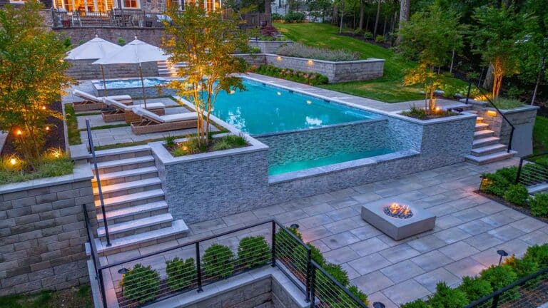 Tiered backyard with illuminated pool, hot tub, fire pit, and stairs at dusk. By Harmony Design Group.