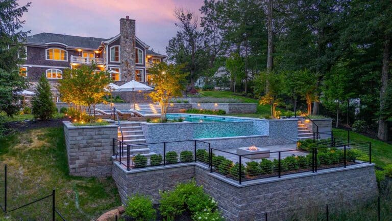 Multi-tiered backyard with illuminated pool, fire pit, and retaining walls, overlooked by a large house at dusk. By Harmony Design Group.