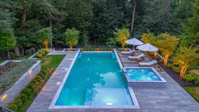 Rectangular pool and square hot tub with illuminated turquoise water, surrounded by gray pavers, lounge chairs, and lush, lit trees by Harmony Design Group.