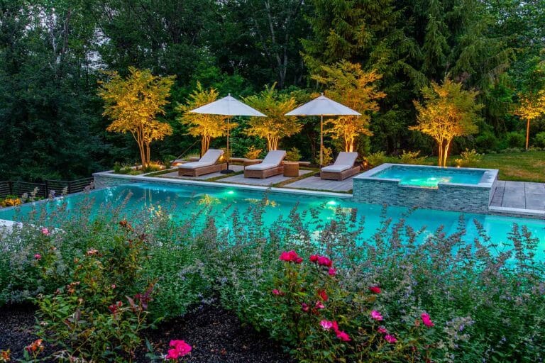 Illuminated aqua swimming pool and hot tub by lounge chairs, umbrellas, and spotlight-lit trees at dusk by Harmony Design Group.